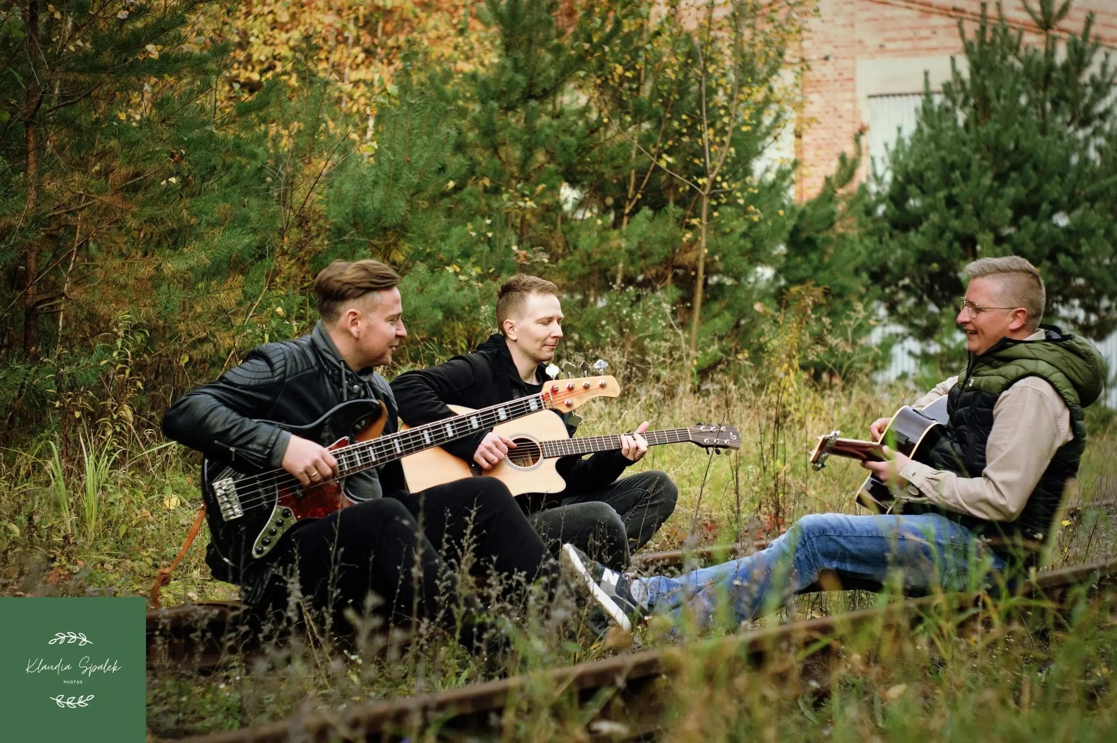 Trzech mężczyzn z gitarami siedzi na nieużywanych torach kolejowych w wysokiej trawie, w tle widać drzewa iglaste i budynek z cegły.
