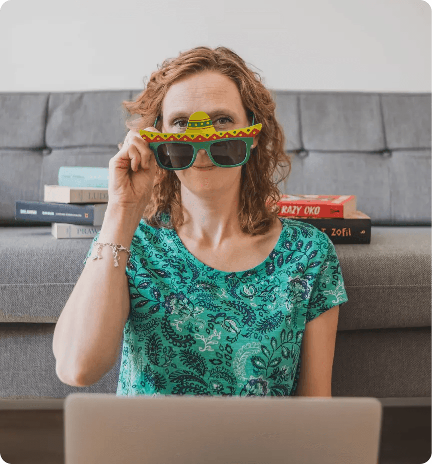 Kamila w zielonych okularach, które przypominają sombrero, przed nią znajduje się laptop, a za nią szara kanapa, na której znajdują się książki.