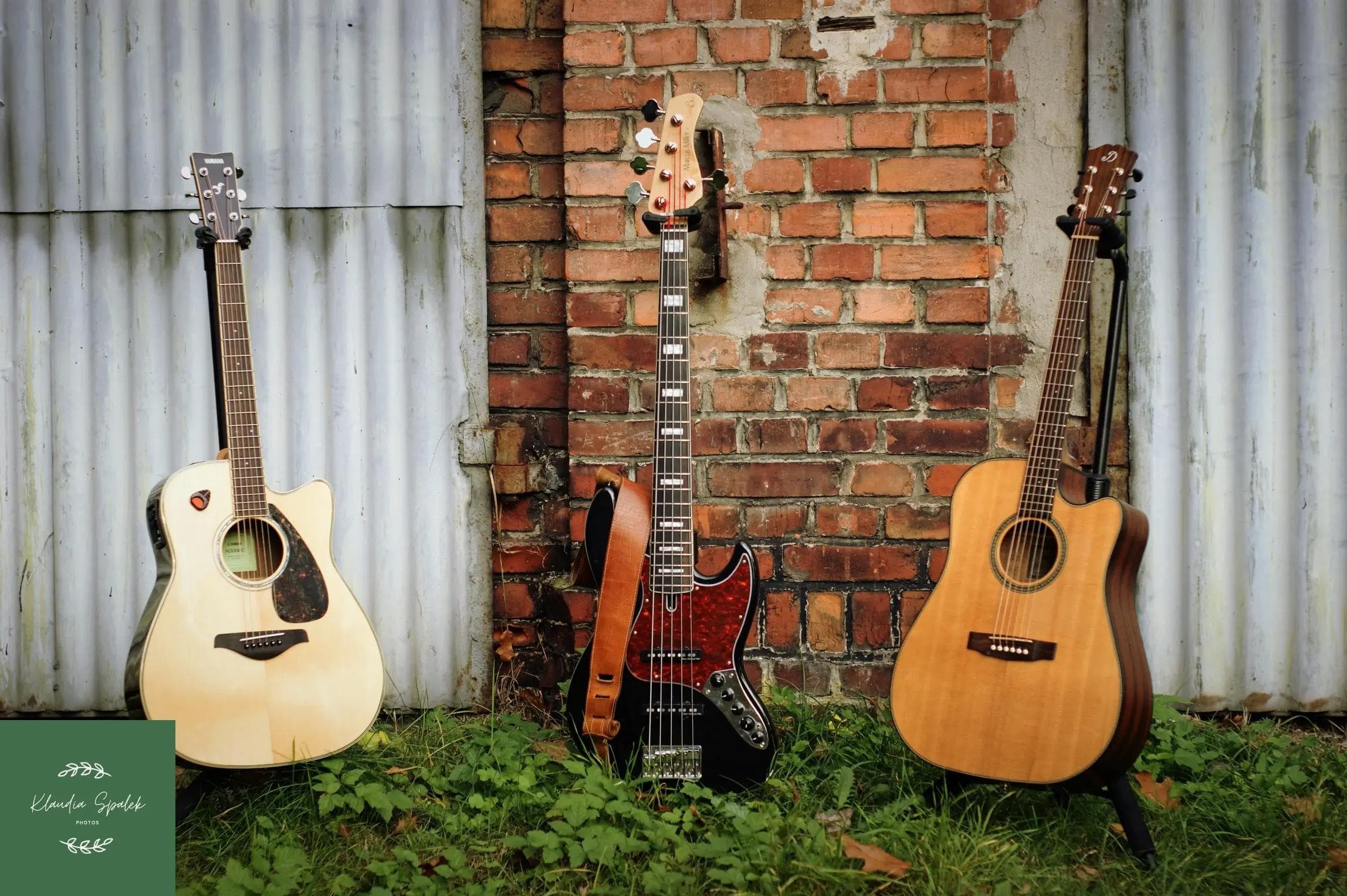 Trzy gitary, w tym dwie klasyczne i jedna elektryczna, stoją na trawie oparte o ceglaną ścianę.