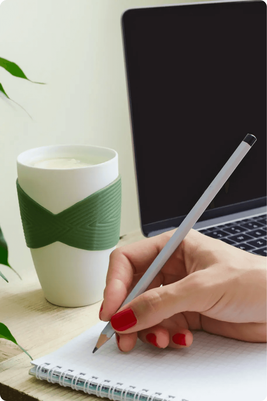 Kobieta trzyma w dłoni ołówek i pisze w notatniku, po prawej stronie znajduje się laptop, a po lewej kubek z napojem.